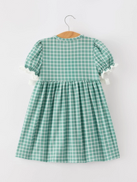 A green and white gingham check dress with short puff sleeves and white ruffle trim, hanging on a wooden hanger against a plain light gray background.