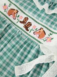 Close-up of a green and white gingham dress with white lace trim and a smocked western-themed band featuring embroidered horses, cowboy boots, and pink flowers.
