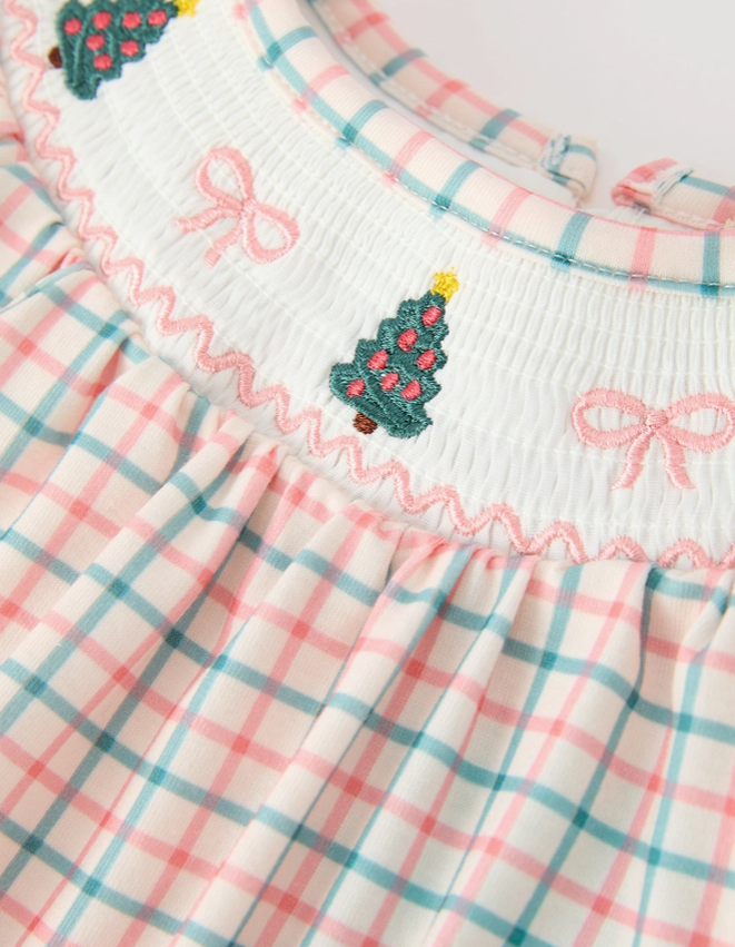 Close-up of a children's dress with a white smocked bodice featuring embroidered pink bows and green Christmas trees, paired with a pink and teal plaid skirt.