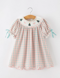 A toddler girl's short-sleeve dress with a pink, green, and white gingham pattern, featuring a white embroidered yoke with pine trees and bows, and light blue ribbon ties on the sleeves, hanging on a wooden hanger against a white background.