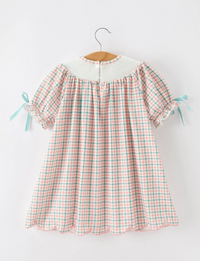 A short-sleeve baby dress with a pink and green gingham pattern, featuring a solid white collar and light blue ribbon ties on the sleeves, hanging on a wooden hanger against a white wall.