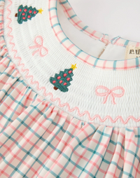 Close-up of a children's dress with a white smocked yoke featuring embroidered pink bows and green Christmas trees, paired with a pink, white, and teal plaid skirt.