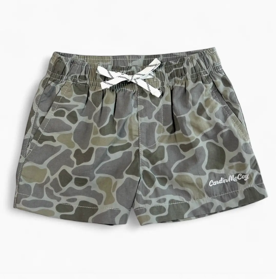A pair of children's camouflage print shorts in shades of gray, olive green, and beige, featuring an elastic waistband with a white bow and a white embroidered 'Cardin McCoy' logo on the lower left leg.