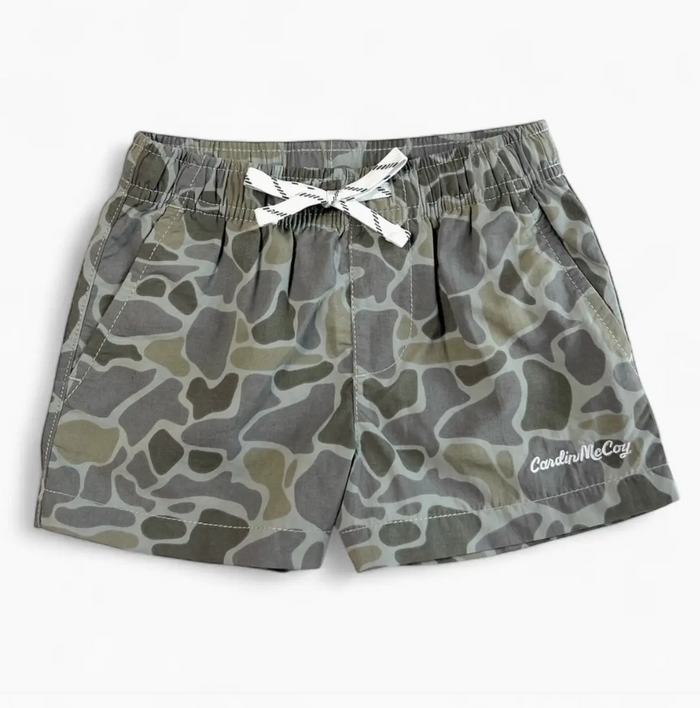 A pair of children's camouflage print shorts in shades of gray, olive green, and beige, featuring an elastic waistband with a white bow and a white embroidered 'Cardin McCoy' logo on the lower left leg.